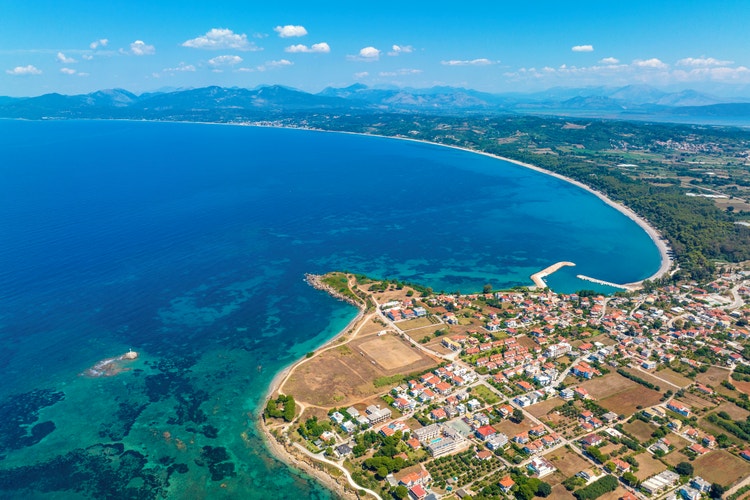Aerial view of Mytikas, featuring a vibrant blue sea, sandy beach, and a scenic coastal town with lush greenery nearby.