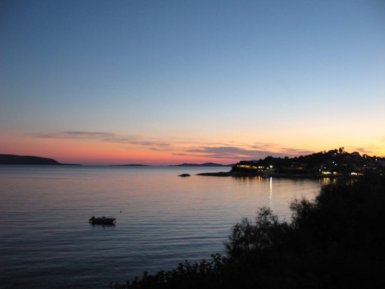 Sunset over Finikounda, Greece, with calm waters reflecting hues of pink and orange, highlighting the coastal village's lights.