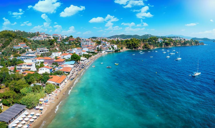 A picturesque seaside view of Megáli Ammos featuring colorful houses, a sandy beach, and sailboats on clear blue waters.