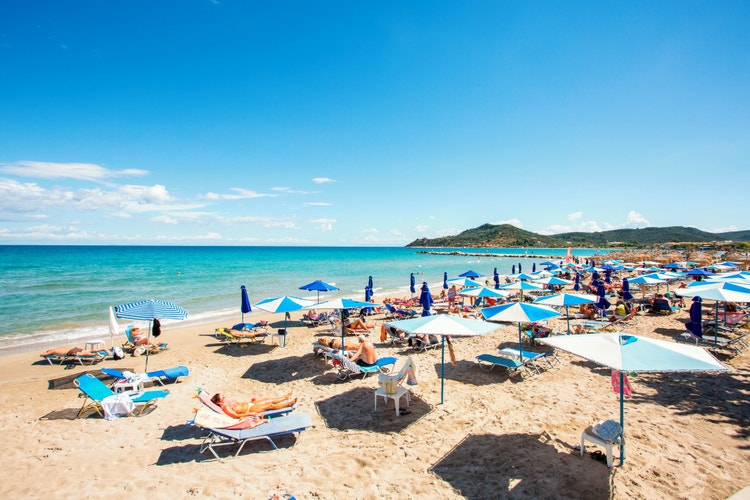 A sunny beach at Alykes, with sunbathers under blue and white umbrellas, clear blue sea and green hills in the background.