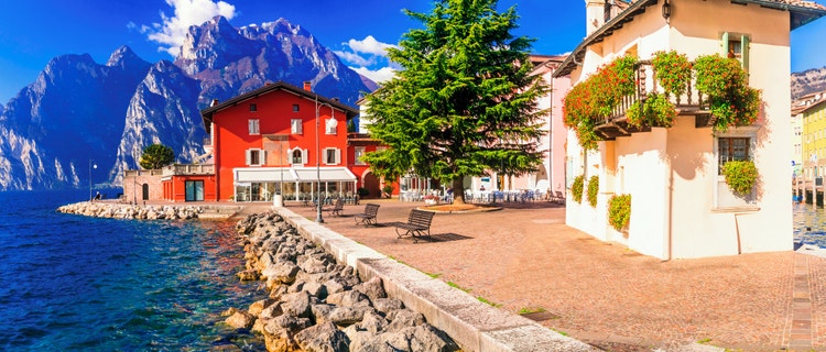 A waterfront promenade in Torbole sul Garda, with red buildings and a sunny atmosphere, with the mountains in the background.