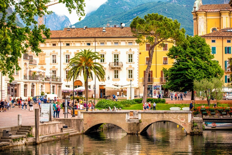 A lively street scene in Riva, featuring palm trees, historic buildings, and people enjoying outdoor cafes and shops.