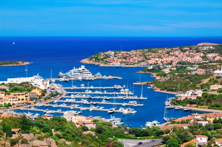 Aerial view of Porto Cervo featuring a marina filled with yachts, colorful buildings, and lush greenery against a blue sea backdrop.