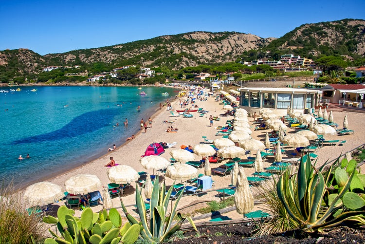 Baia Sardinia beach with sunbathers, umbrellas, and clear blue water. Tourists enjoy swimming and relaxing on the sand.