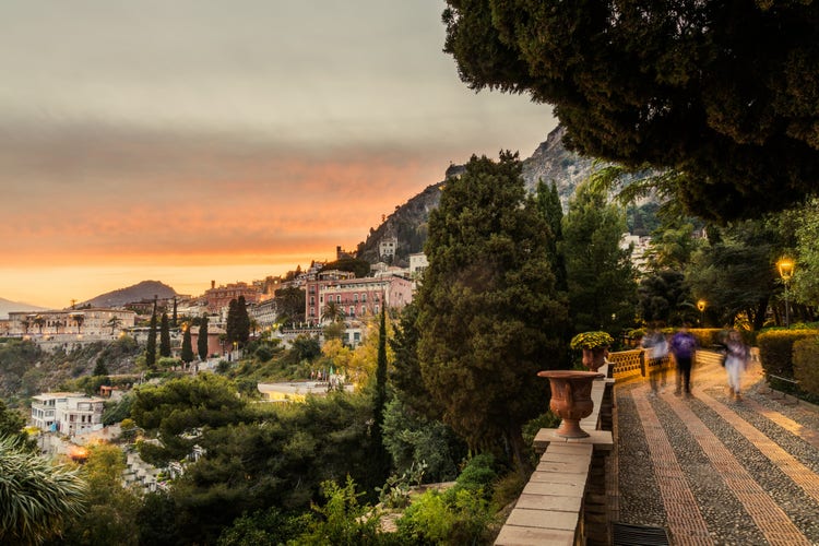 Sunset over Taormina, featuring hills, historic buildings, and lush greenery, with people walking along a charming path.