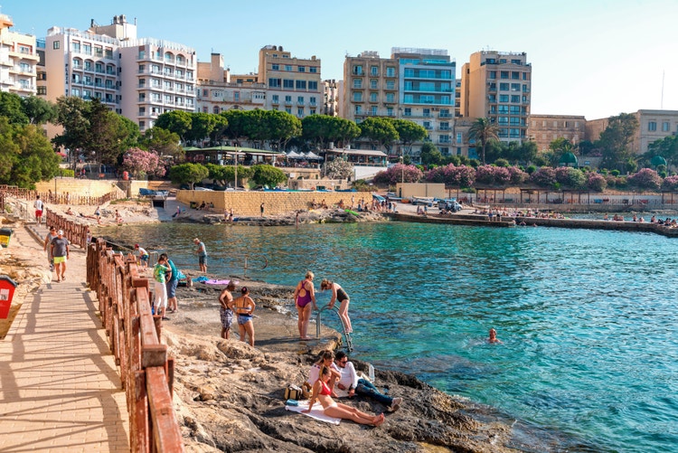 Sliema,people are swimming, sunbathing, and enjoying the sunny waterfront. Lush greenery, pink flowers and restaurants in the background.