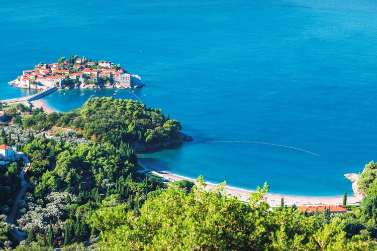 Aerial view of Sveti Stefan, framed by lush greenery and blue waters, with boats in the background.