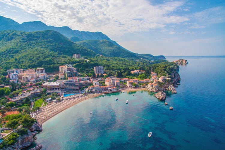 Aerial view of the seaside village of Petrovac, showing coastal cliffs, red-roofed buildings and clear blue waters.