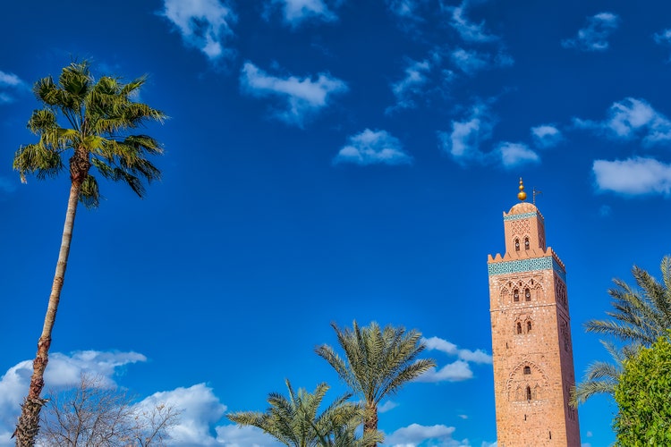 A vibrant blue sky frames the iconic Koutoubia Mosque tower in Marrakech, with palm trees gently swaying in the foreground.