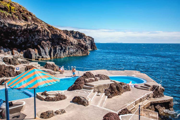 A serene ocean view from Canico, featuring a pool, rocky coastline, and people enjoying the sun near the water.