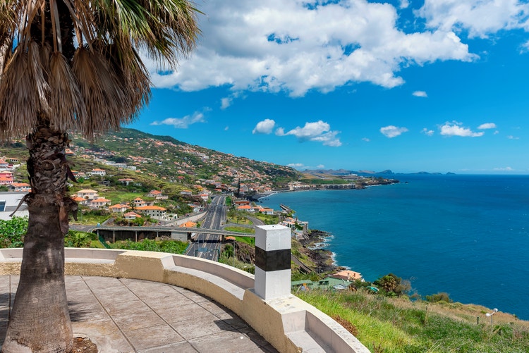 A scenic view of Santa Cruz, Madeira, with lush green hills, houses, a coastal road and the blue Atlantic Ocean.