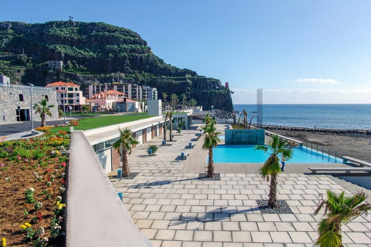 A scenic view of Ribeira Brava featuring palm trees, a swimming pool, vibrant gardens, and a coastline with the ocean in the background.