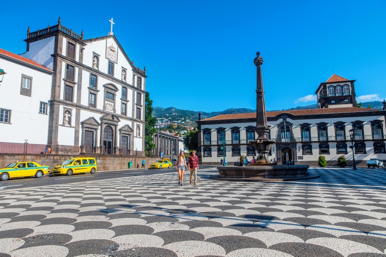 A beautiful view of a square in Funchal on a sunny day, showing a fountain facing a white church and yellow taxis parked.