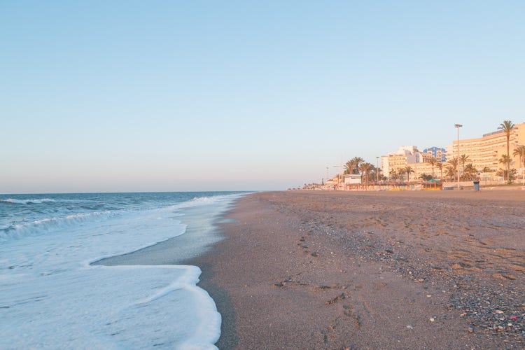 Roquetas de Mar beach at sunset, featuring gentle waves and a sandy shoreline with buildings and palm trees in the background.