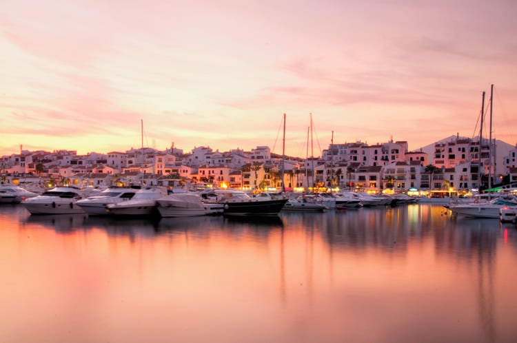 Sunset view of Puerto Banus marina, featuring numerous yachts and upscale buildings reflecting on the calm water.