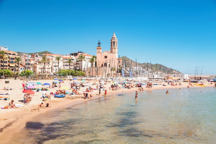 Sitges beach, with sunbathers, colourful parasols and palm trees against a backdrop of boats and charming architecture.