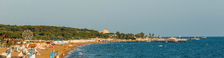 A sunny beach scene in Sorgun with sunbathers, a pier, and a ferris wheel amidst greenery and calm turquoise waters.