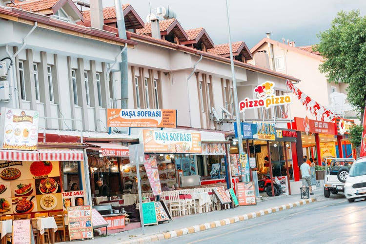 A street with various restaurants, including Paşa Sofrası and Pizza Pizza, in Ovacik, with colorful signage and decorations.