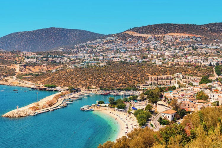 A scenic view of Kalkan, Turkey, showcasing coastal boats, hillside homes, and rural areas under a clear blue sky.