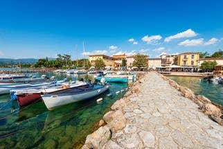 Stone pier leads to colourful boats in clear, green-blue water. Vibrant buildings line the peaceful harbour in Cisano.