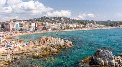 A panoramic view of Lloret de Mar beach with sunbathers, umbrellas, and clear waters, framed by seaside buildings and hills.