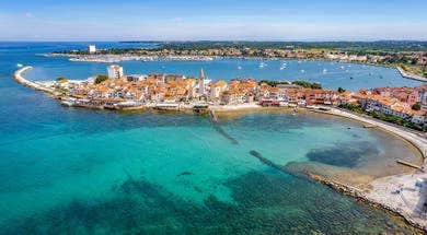 Aerial view of a Umag with terracotta rooftops, surrounded by clear turquoise waters and a marina with small boats. The sky is clear and blue.