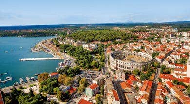 A tranquil beach in Pula, featuring clear blue waters, rocky cliffs, sunbathers, and swimmers enjoying the sunny day.
