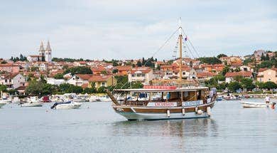 Boat named Excursion Zlatni Rat sailing on calm water in Medulin with red-roofed houses and church towers in the background.