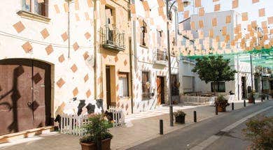 A sunny street with patterned facades, white picket fences, and a cow cut-out. Hanging orange and green banners in Pineda de Mar on Spain’s Costa Barcelona