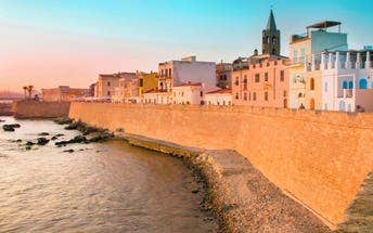 Coastal view of Alghero, Sardinia, shows pastel-coloured buildings atop a historic stone wall at sunset, overlooking a serene, rocky shoreline.
