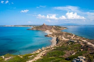 A scenic coastal landscape of Oristano with a narrow land strip extending into a calm turquoise sea. A small stone tower sits on the land under a bright blue sky.