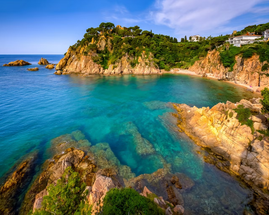 Scenic coastal view of a turquoise bay surrounded by rocky cliffs and lush greenery, with scattered houses in Blanes on the Costa Brava.